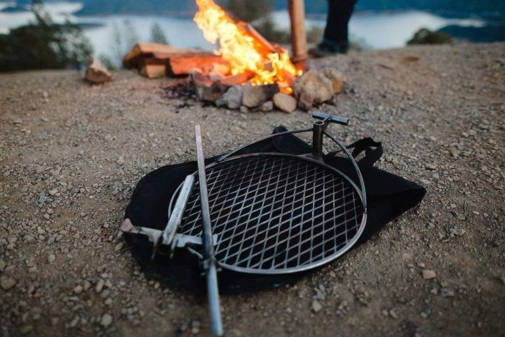 Campfire by a river with the Outpost grilling system in front of it laying on the bag.