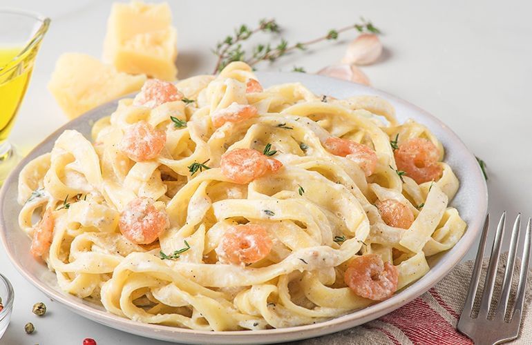macarrão tipo espaguete com molho branco e camarão em cima de uma mesa branca com garfo ao lado,