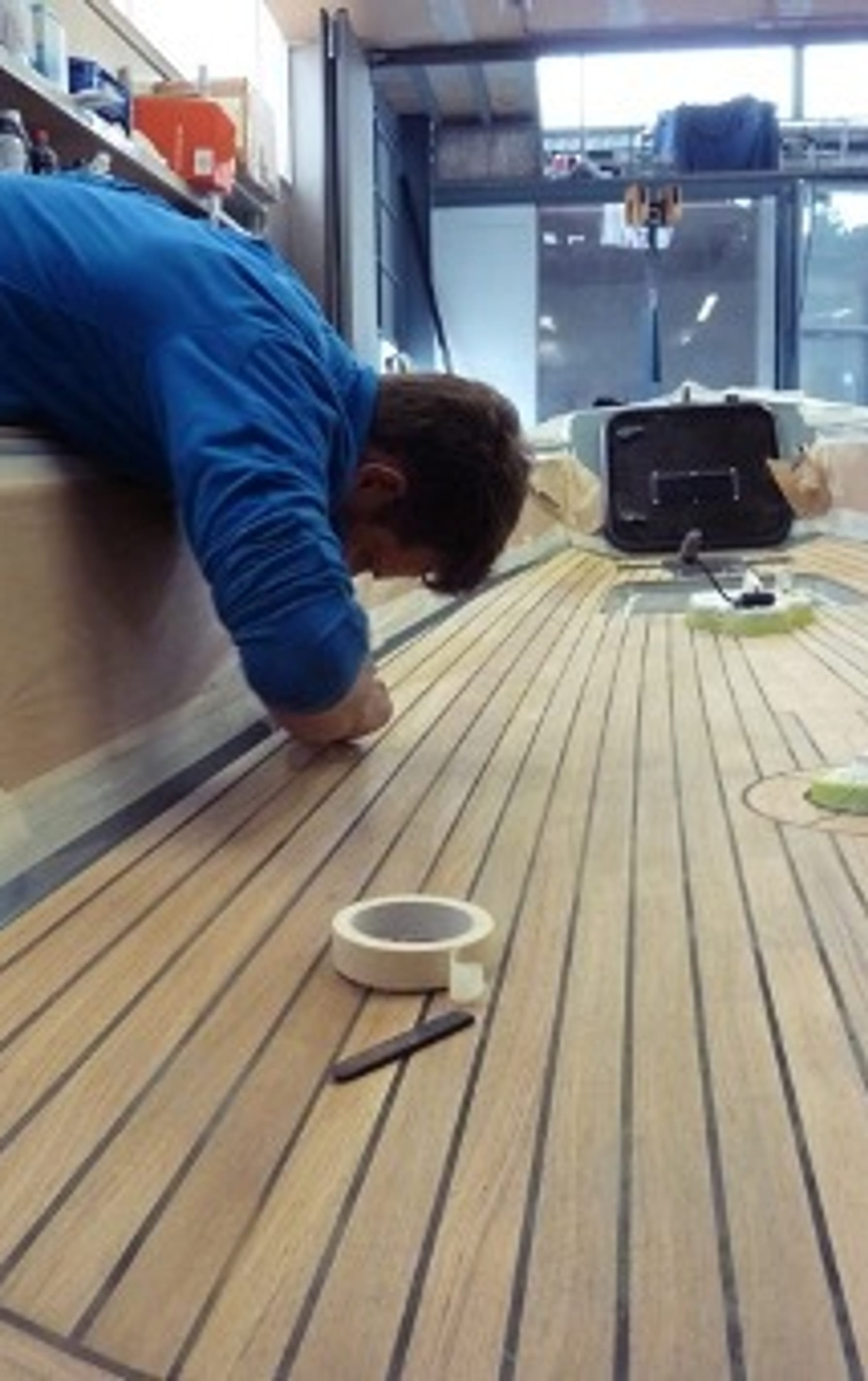 Person in blauer Arbeitskleidung montiert sorgfältig das Holzdeck eines Esse Segelbootes in unserer Werkstatt, mit Werkzeugen und Abdeckmaterial in der Umgebung.