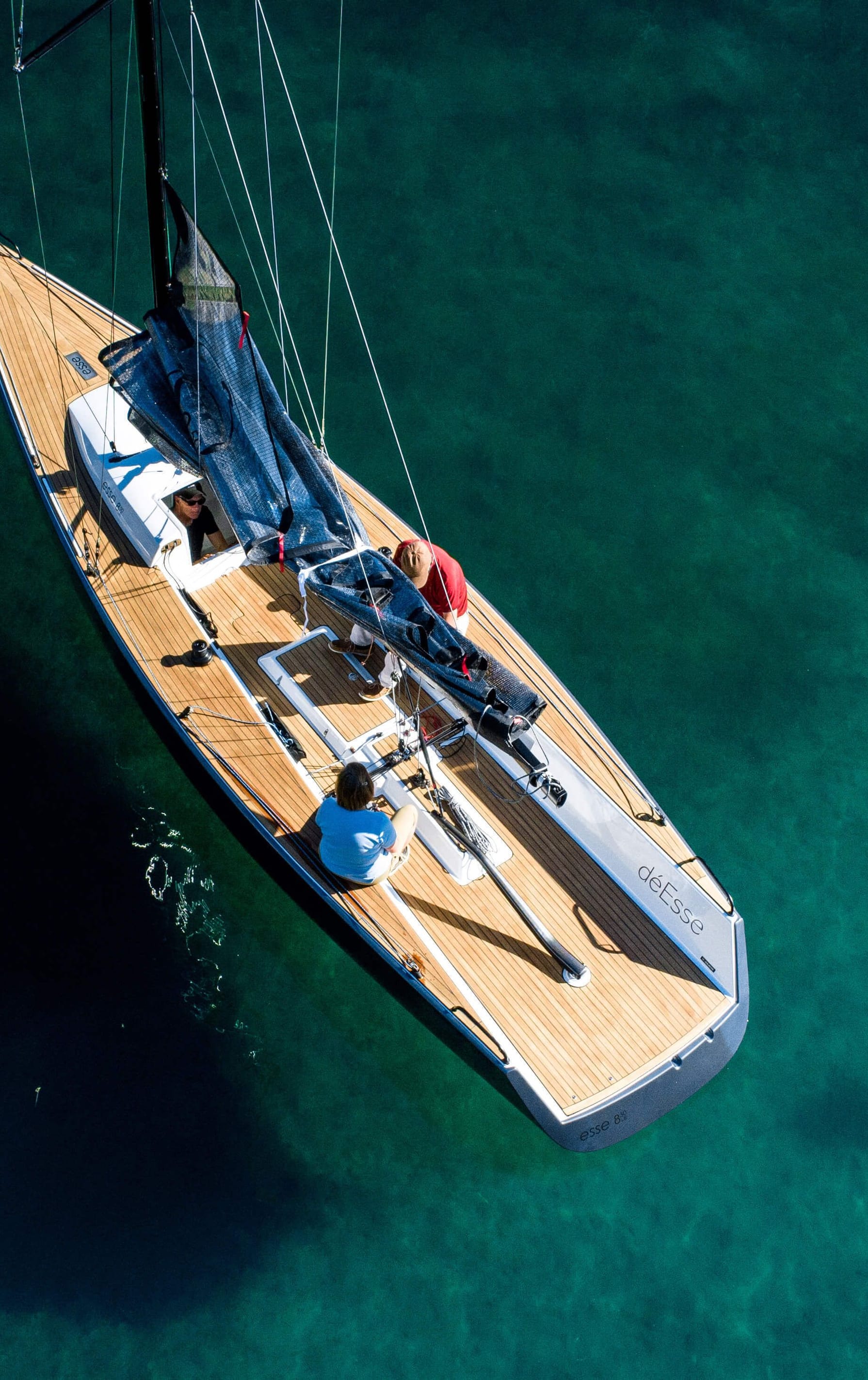 Luftaufnahme eines eleganten Segelboots mit Holzdeck im klaren, türkisfarbenen Wasser, neben einer roten Boje, mit mehreren Personen an Bord, die das Boot bedienen oder entspannen.