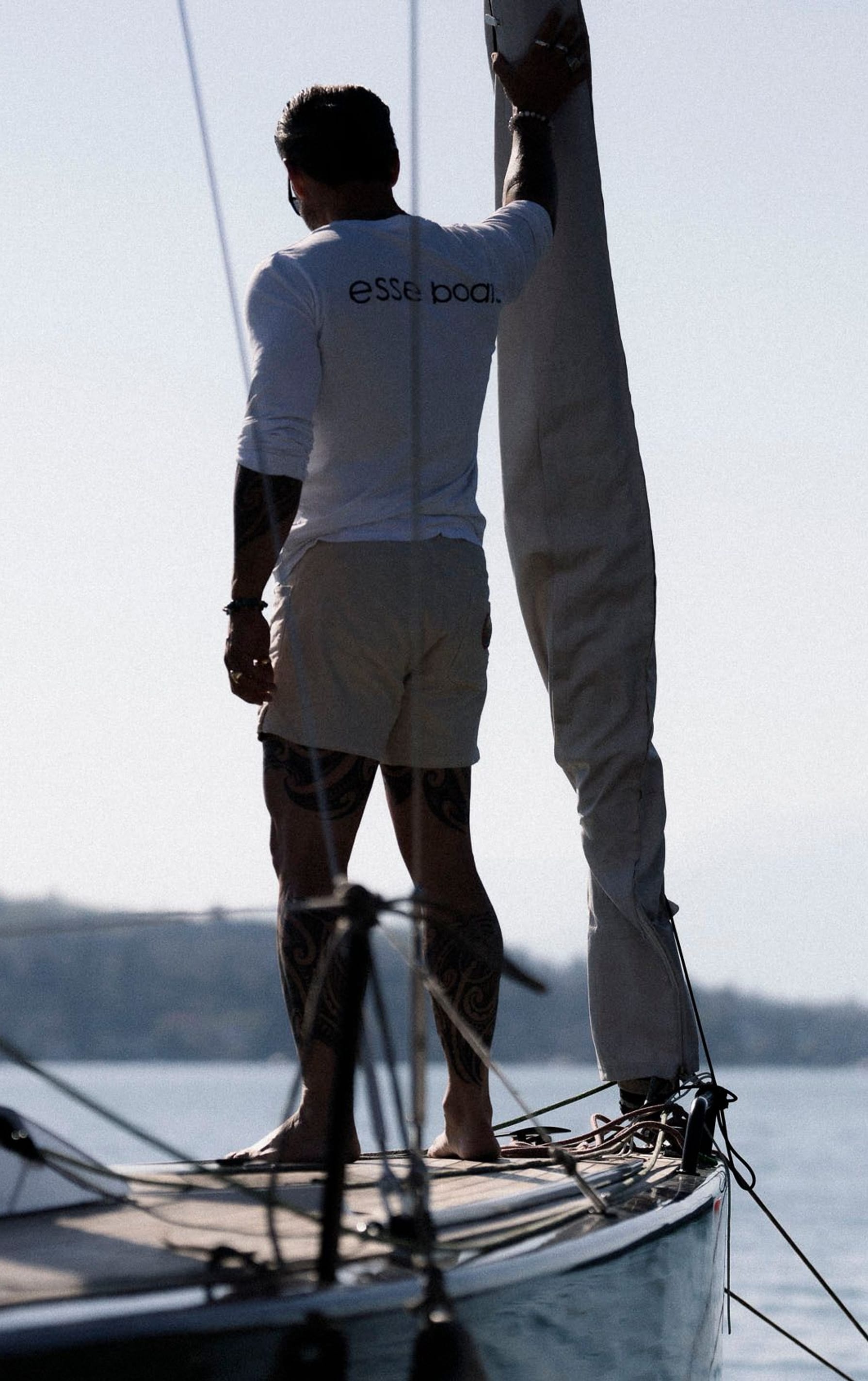 Silhouette eines Seglers, der auf dem Vorderdeck eines Segelboots steht und das Segel bedient, mit dem Logo 'esse boats' auf seinem weißen Shirt, gegen einen diffus beleuchteten Hintergrund mit Wasser und anderen Booten.