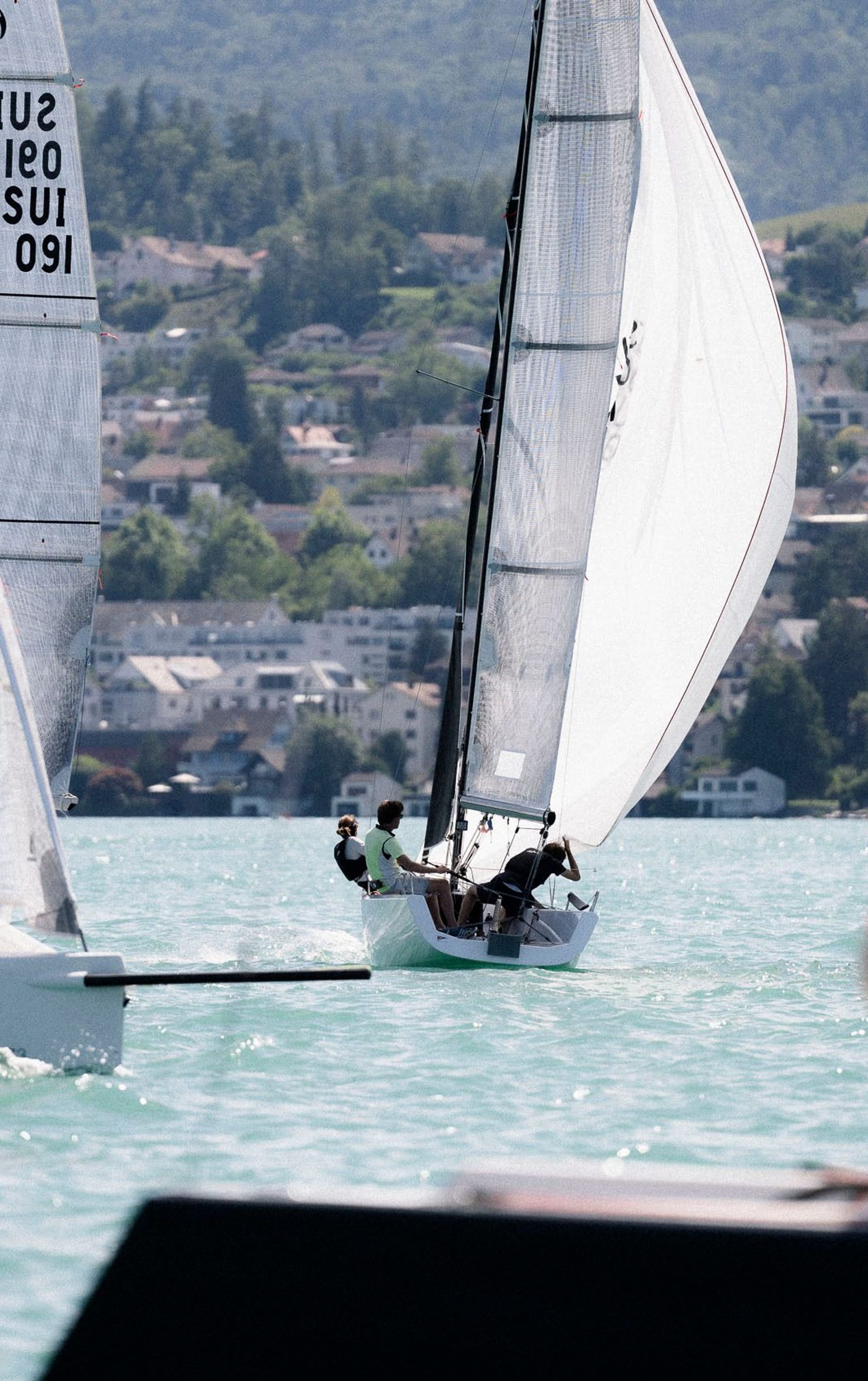 Regattasegler auf Segelbooten konzentrieren sich auf das Rennen auf einem blauen See vor der Kulisse einer Stadt, mit klarem Fokus auf das Boot im Vordergrund und der URL esseboats.ch auf den Segeln.