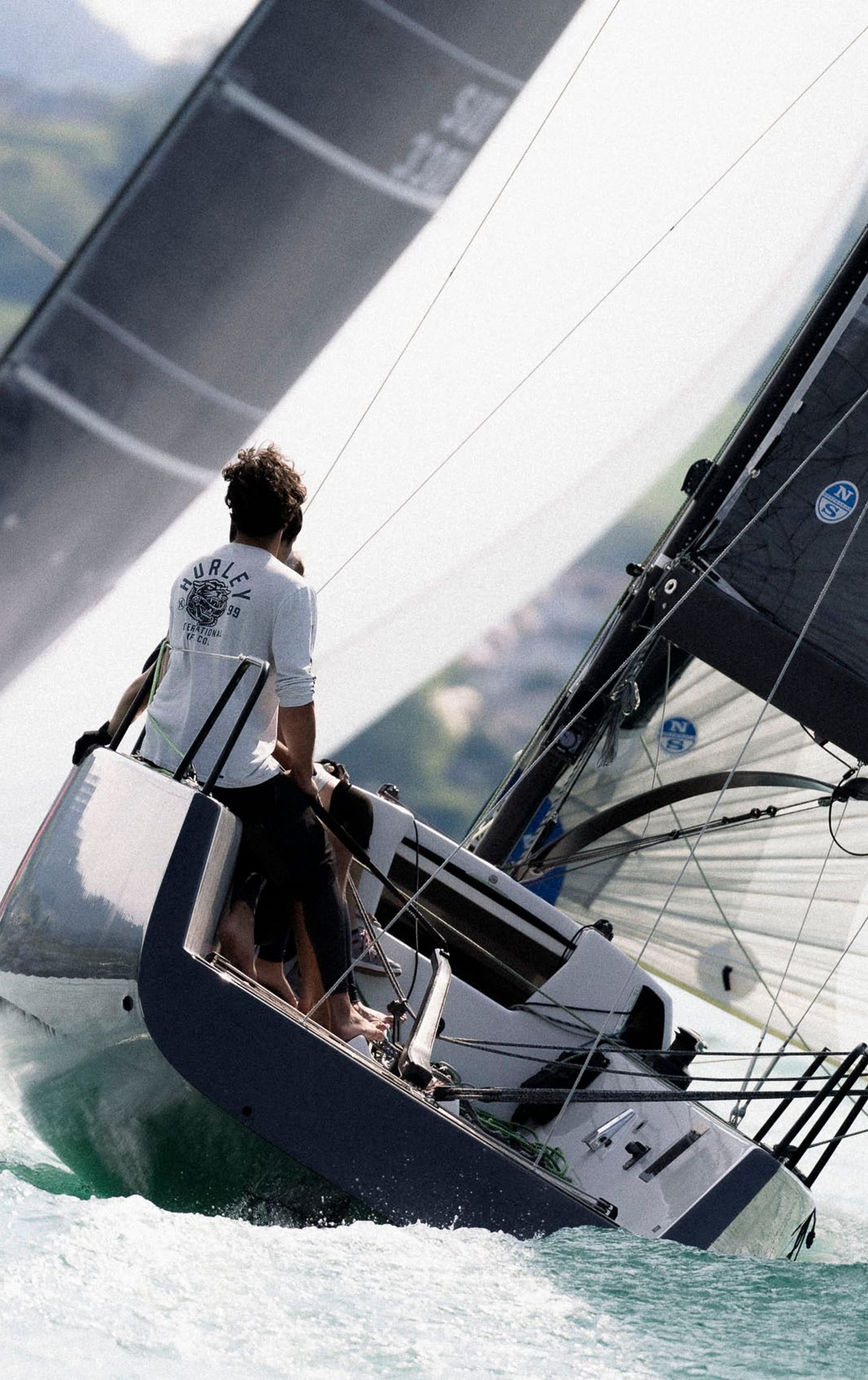 Zwei Segelboote mit aufgeblähten schwarzen Segeln rasen über das Wasser, mit Crewmitgliedern, die sich konzentriert auf ihre Aufgaben fokussieren; im Hintergrund ist eine grüne Landschaft zu sehen. Weitere Informationen finden Sie auf esseboats.ch.