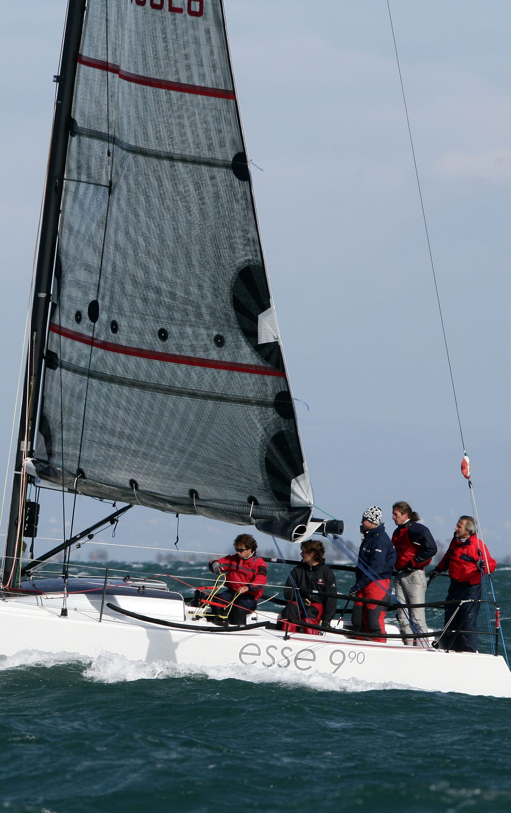 Ein Segelteam in roten und schwarzen Jacken bedient ein Segelboot der Klasse 'Esse 990' auf welligem Wasser. Weitere Informationen finden Sie auf esseboats.ch.
