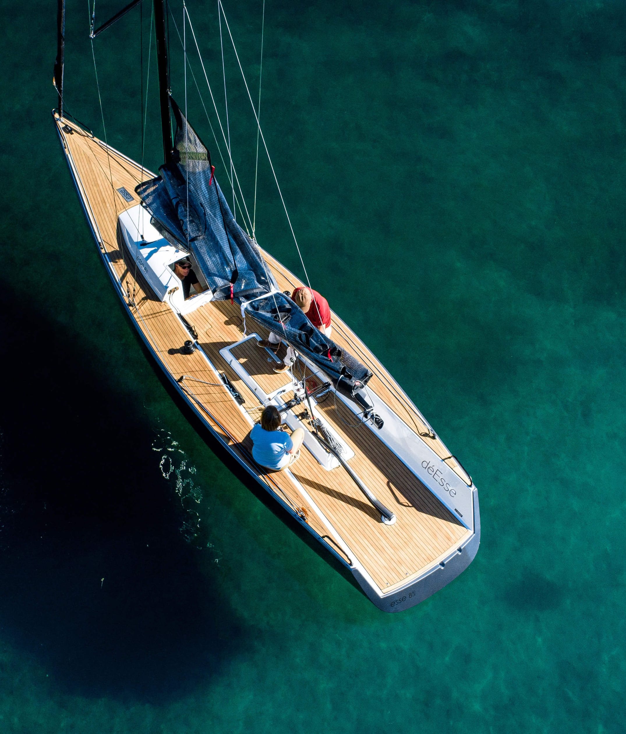 Luftaufnahme eines eleganten Segelboots mit Holzdeck im klaren, türkisfarbenen Wasser, neben einer roten Boje, mit mehreren Personen an Bord, die das Boot bedienen oder entspannen.
