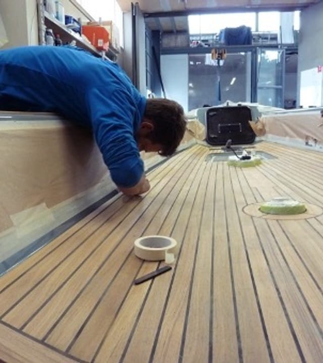 Person in blauer Arbeitskleidung montiert sorgfältig das Holzdeck eines Esse Segelbootes in unserer Werkstatt, mit Werkzeugen und Abdeckmaterial in der Umgebung.