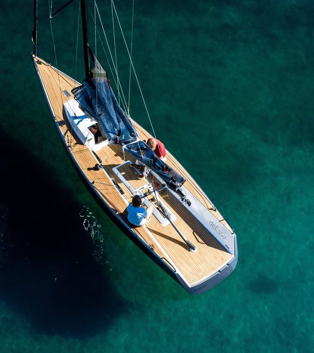 Luftaufnahme eines eleganten Segelboots mit Holzdeck im klaren, türkisfarbenen Wasser, neben einer roten Boje, mit mehreren Personen an Bord, die das Boot bedienen oder entspannen.