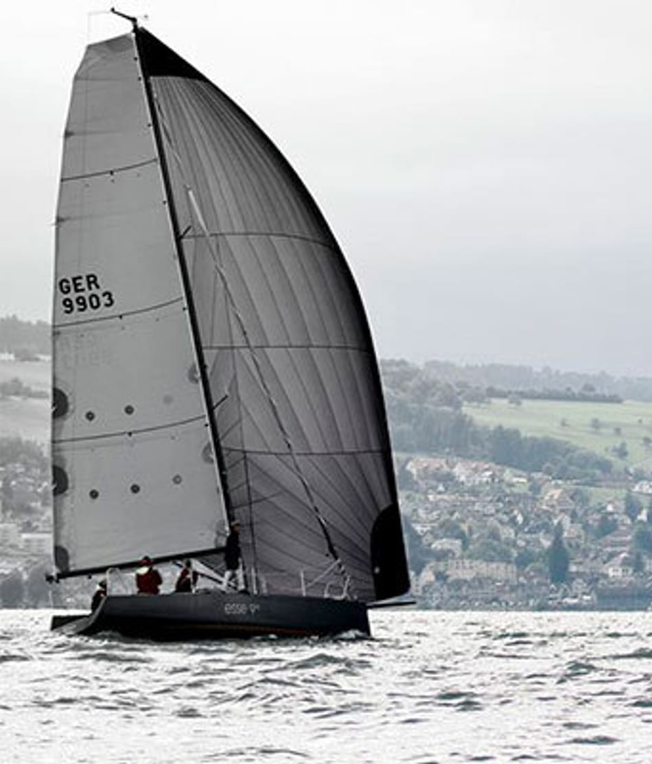 Esseboats esse990 volle segel: Segelboote mit dunklen Segeln auf rauem Gewässer bei einem Segelwettbewerb, mit dem Ufer und grünen Hügeln im Hintergrund. Beschriftung auf den Segeln zeigt 'GER 9903' und 'SUI 99001'. Für weitere Informationen besuchen Sie esseboats.ch.