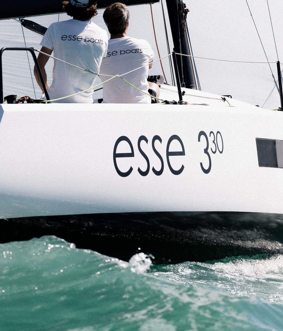 Ein Segelboot der Marke 'esse 330' auf offenem Wasser, fokussiert auf den Heckbereich mit spritzendem Wellenwasser und zwei Personen in Shirts mit der Aufschrift 'esse boats', die sich auf das Segeln konzentrieren.