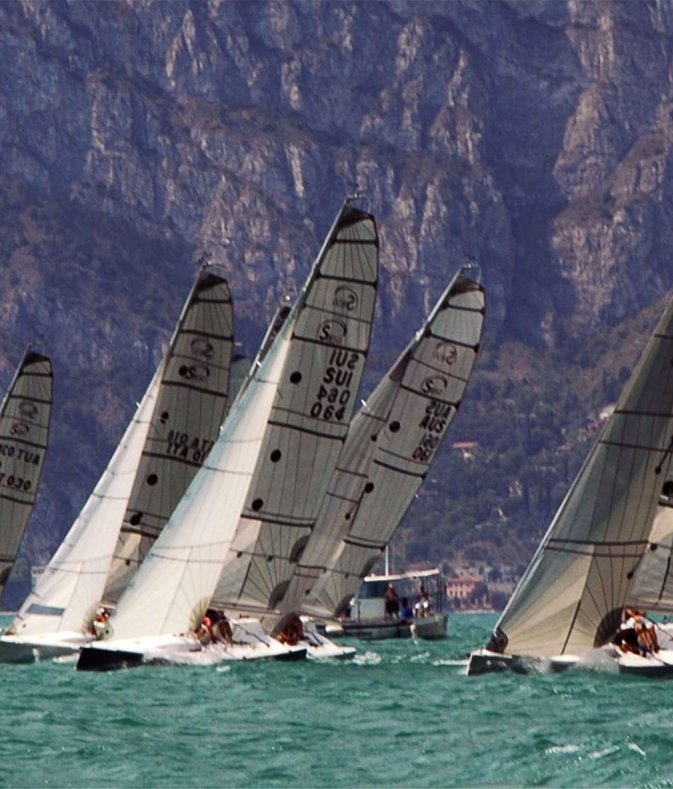 Segelboote der Klasse 'Esse 850' segeln in einer Regattalinie auf dem Gardasee mit dem beeindruckenden Bergmassiv im Hintergrund. Weitere Details finden Sie auf esseboats.ch.