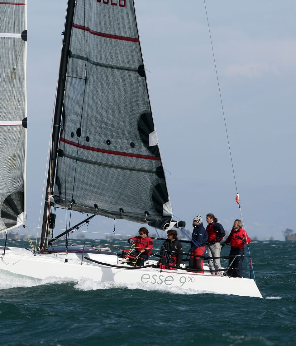 Ein Segelteam in roten und schwarzen Jacken bedient ein Segelboot der Klasse 'Esse 990' auf welligem Wasser. Weitere Informationen finden Sie auf esseboats.ch.