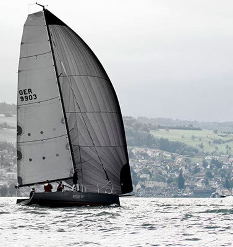 Esseboats esse990 volle segel: Segelboote mit dunklen Segeln auf rauem Gewässer bei einem Segelwettbewerb, mit dem Ufer und grünen Hügeln im Hintergrund. Beschriftung auf den Segeln zeigt 'GER 9903' und 'SUI 99001'. Für weitere Informationen besuchen Sie esseboats.ch.