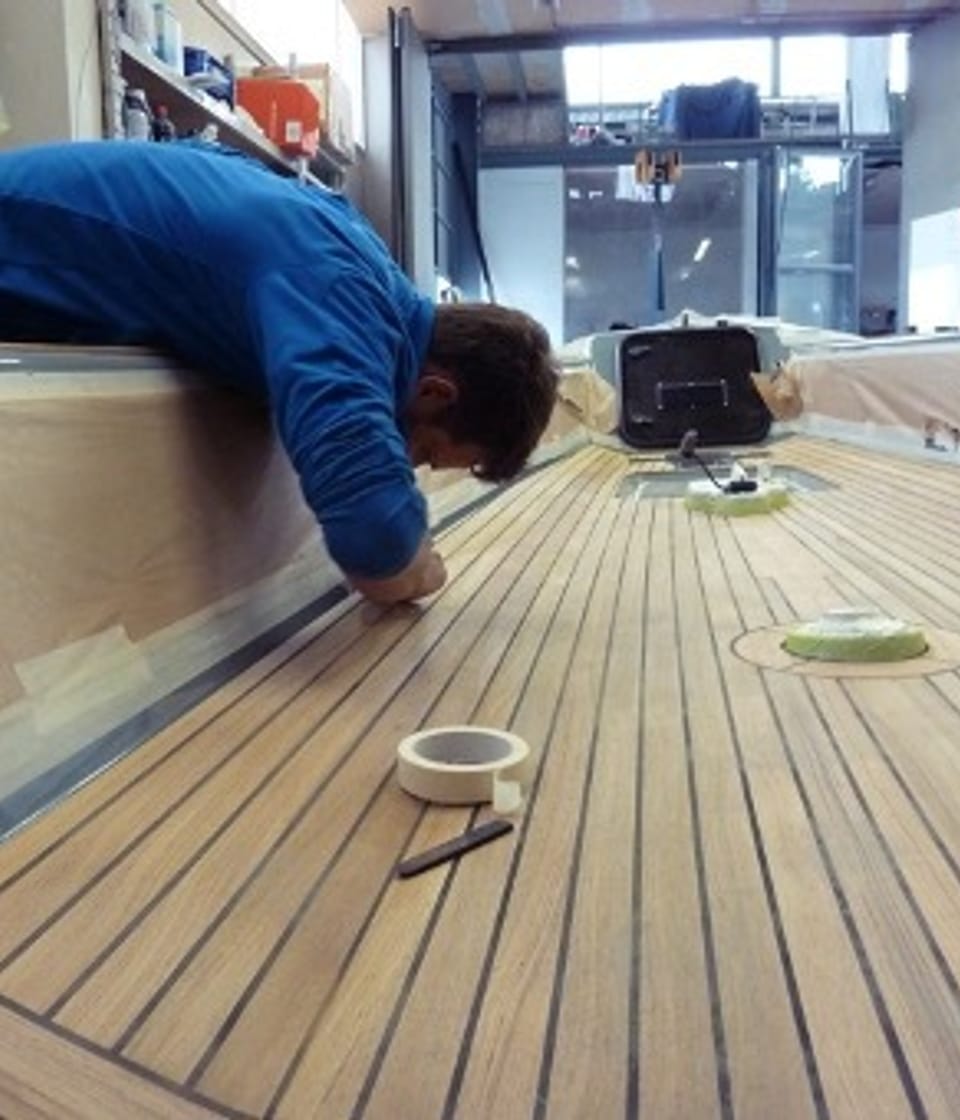 Person in blauer Arbeitskleidung montiert sorgfältig das Holzdeck eines Esse Segelbootes in unserer Werkstatt, mit Werkzeugen und Abdeckmaterial in der Umgebung.