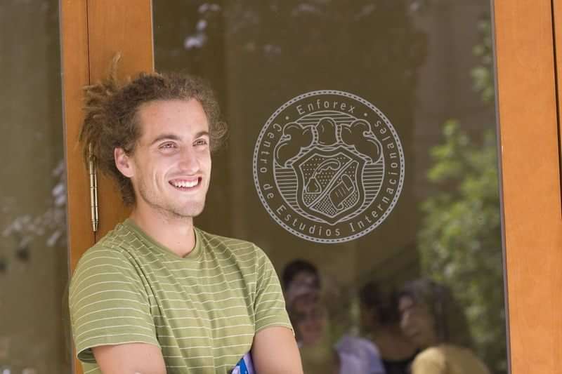 Student bij een taalreisinstelling, glimlachend voor de ingang van het gebouw.