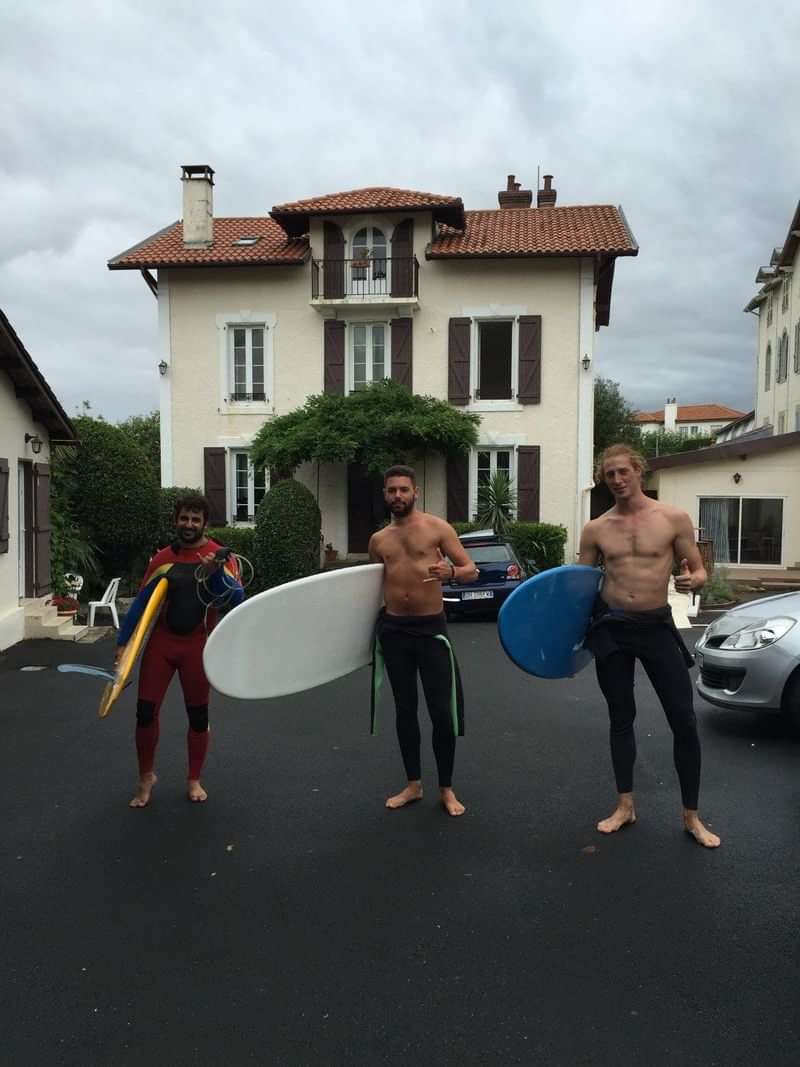 Three surfers standing outside a house, holding surfboards.