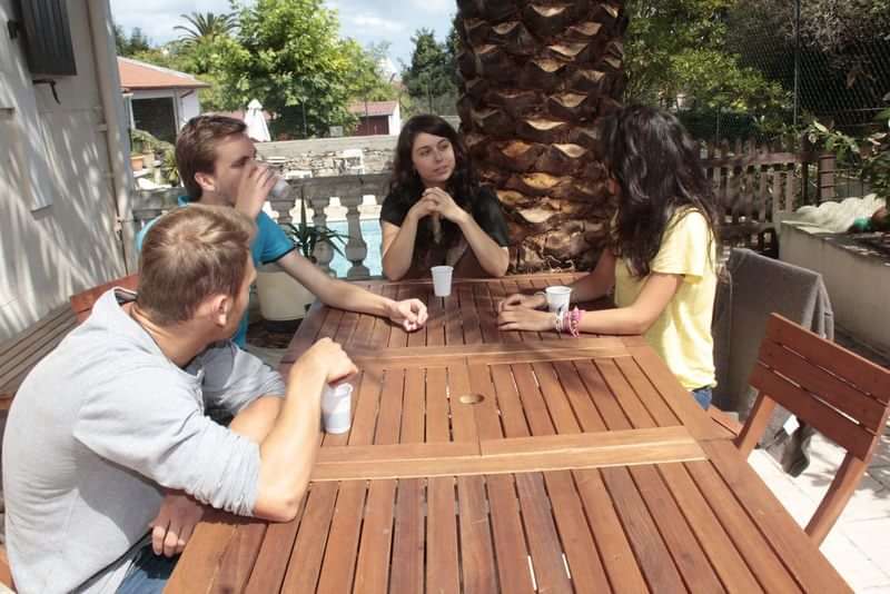 Groep studenten oefent conversatie tijdens een taalreis aan een buitentafel.