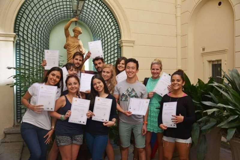 Group of students celebrating language course graduation, holding certificates, outdoor setting.