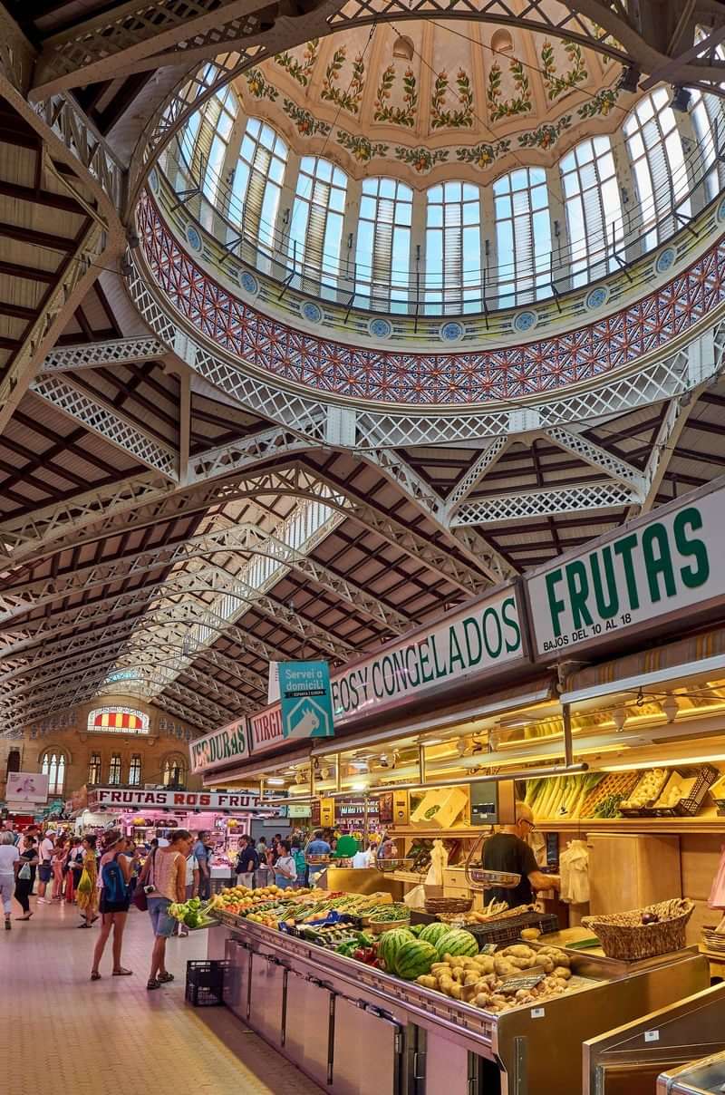 Historic market hall with fresh produce, signage in Spanish, bustling atmosphere, intricate architecture.