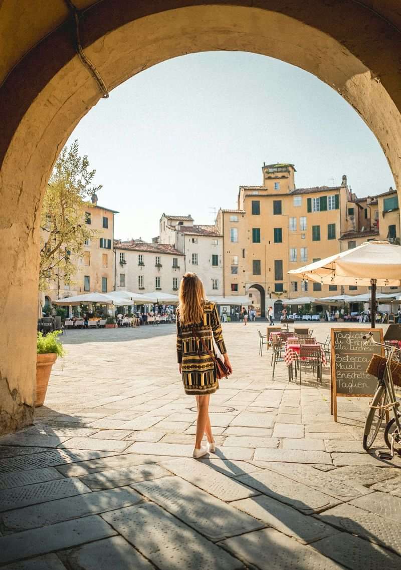 Vrouw wandelt door boog in charmant plein met cafés en restaurants.
