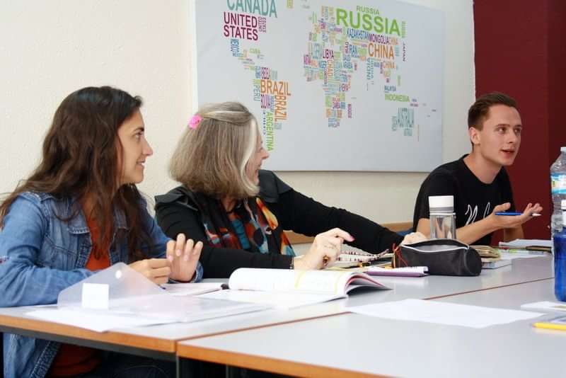 Studenten in een klaslokaal, wereldkaart achter hen, bespreken taalstudie.