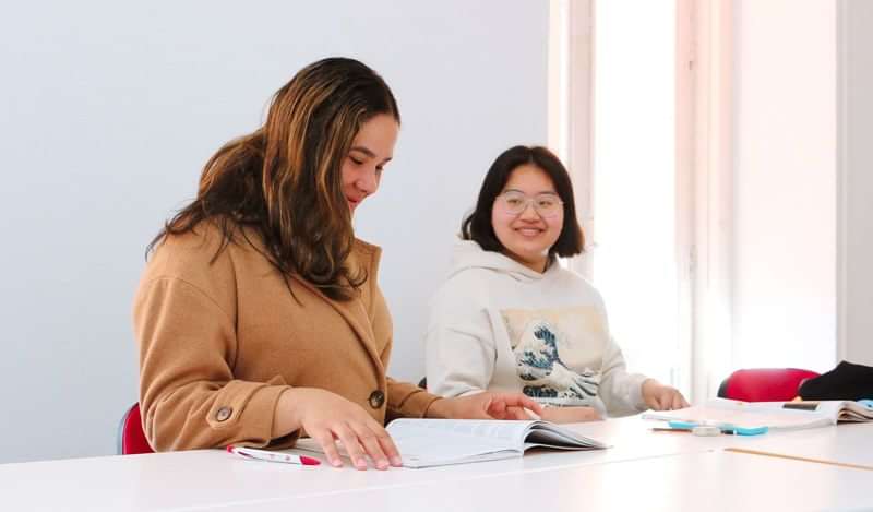 Twee studenten in een taalklas, lezend en glimlachend naar elkaar.