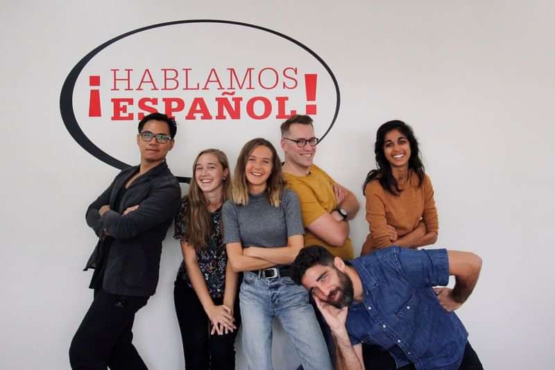 Groep mensen, lachend onder een "Hablamos Español" bord, taalreis sfeer.