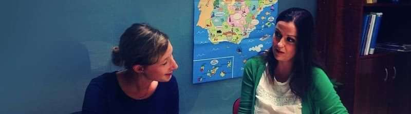 Two women conversing, a map of Spain behind them.