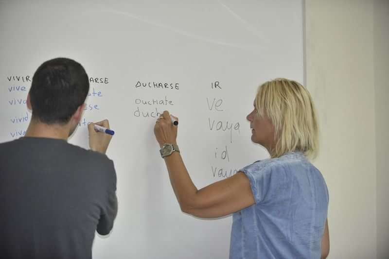 Twee mensen schrijven vervoegingen van werkwoorden op een whiteboard.