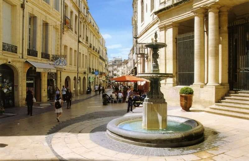 Een levendige straat met een fontein, café en winkels.