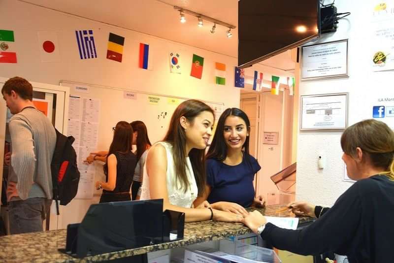 Students at a language school reception desk with international flags overhead.