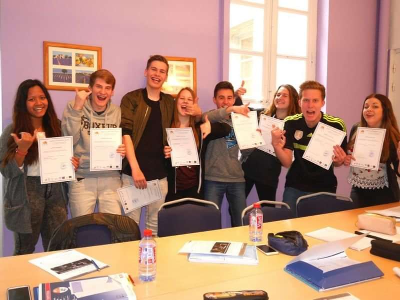 Students celebrating language course completion, holding certificates in a classroom.