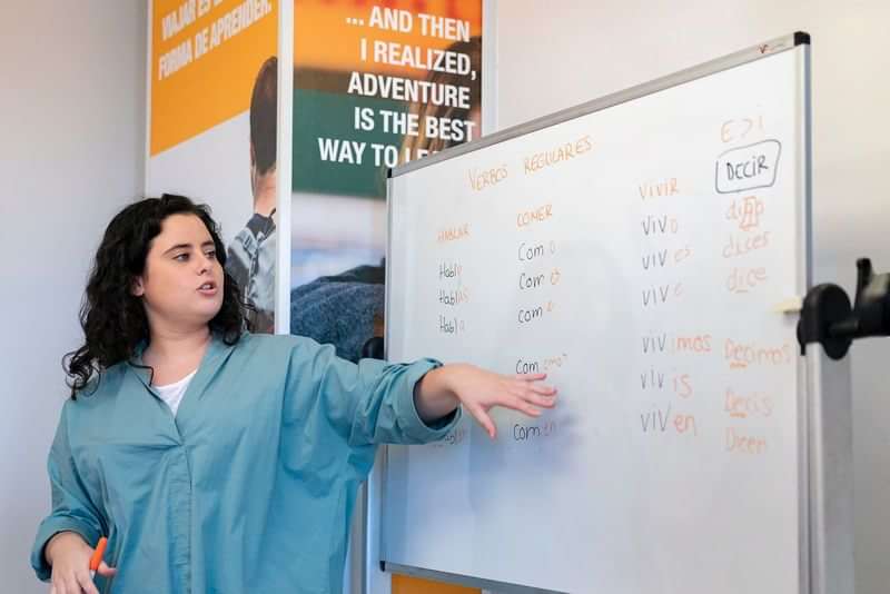 Lerares geeft taalles, wijst naar het whiteboard met vervoegingen.