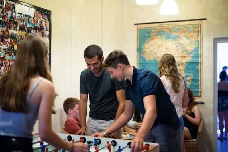 Students socializing, playing foosball, with map in background suggesting travel.