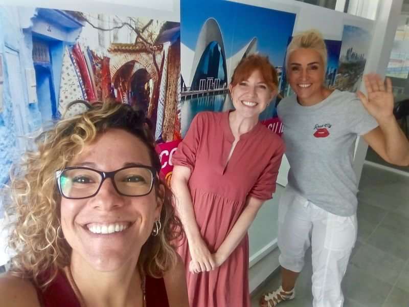 Three women smiling in front of international travel posters.