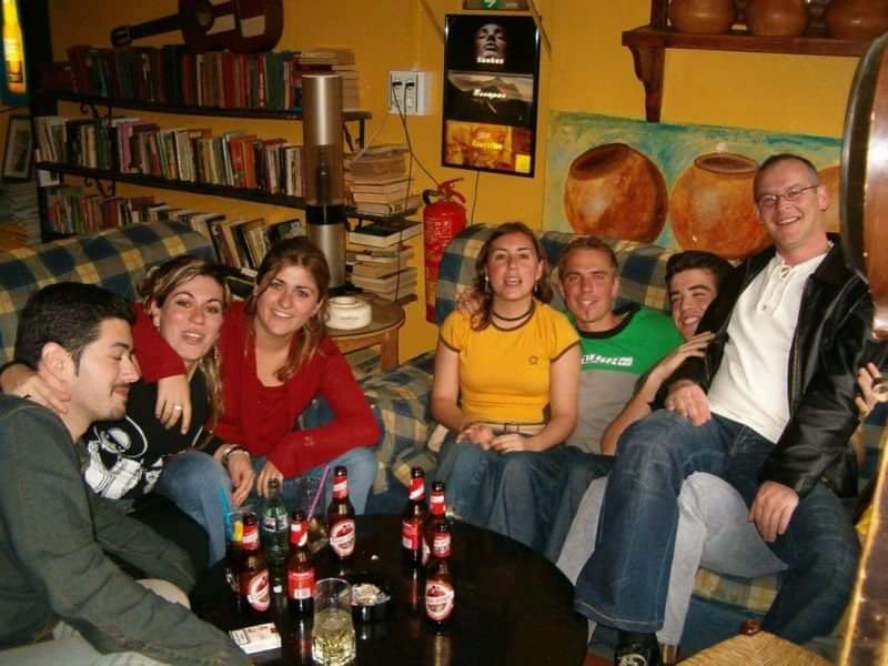 Groep mensen ontspannen, lachen, drinken bier, boekenplanken op achtergrond.