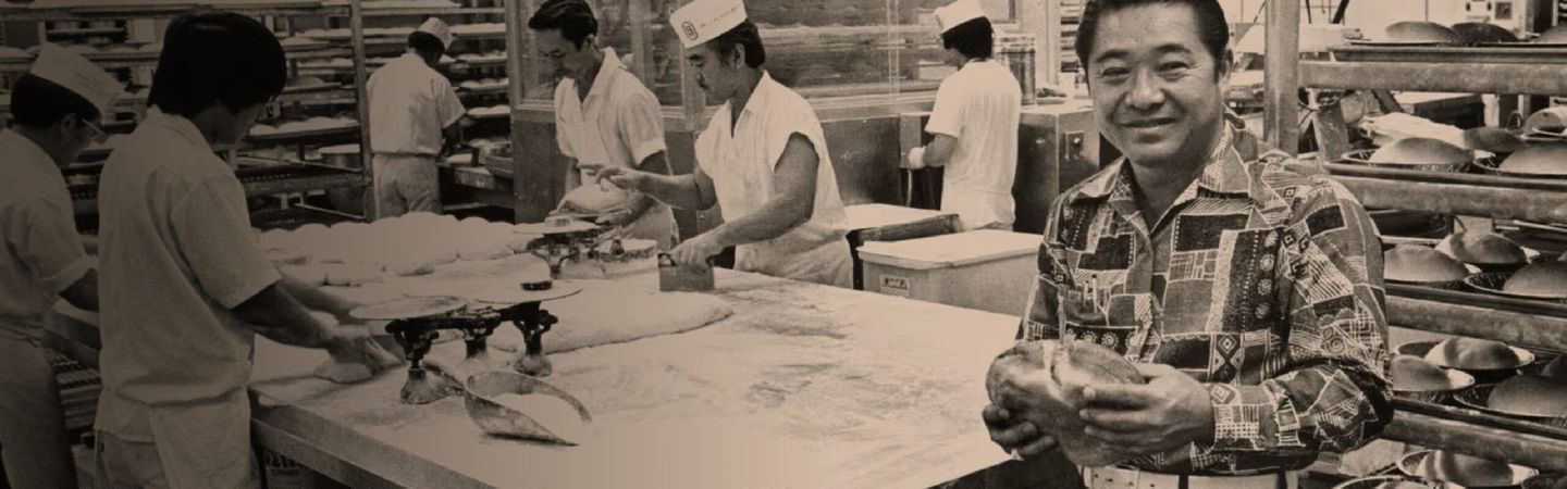 Robert R. Taira in his original bakery factory holding his Original Hawaiian Sweet bread loaf (sepia photo)