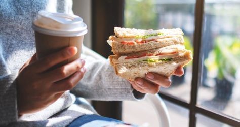 Individual holding a sandwich and a coffee in front of a window							