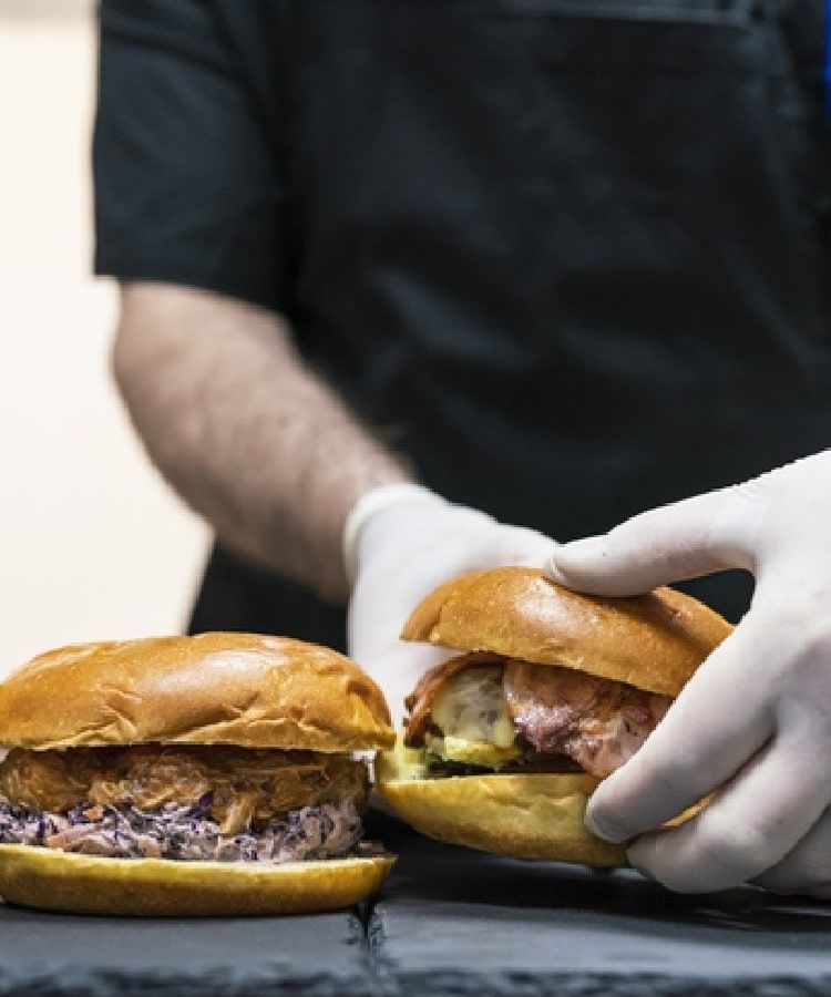 Chef in white gloves holding one of three burgers on a grey slate