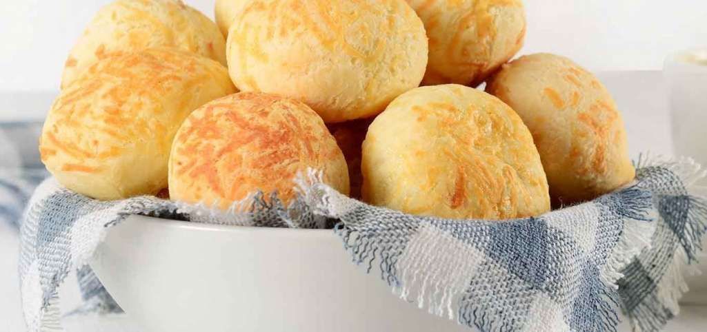 Um uma tigela branca com um pano de prato xedrez de branco e azul claro vemos vários pães de queijo com a textura granulado e cor amarelo claro e detalhes em laranja.