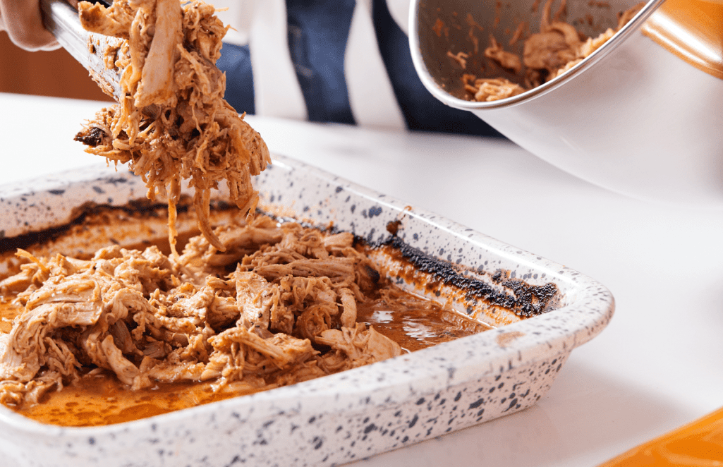 Tigela de cerâmica branca com bolinhas pretas com frango desfiado temperado na cor marrom. Alguém pega com um pegador mais frango de uma panela para por na tigela.