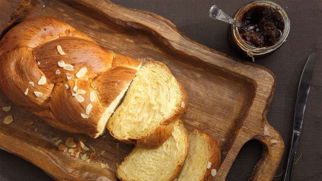 Pão doce alem]ao trançado em cima de uma tábua de cortes de madeira cortado em algumas fatias. Sua cor por fora é marrom brilhante e por dentro um bege clarinho. Ao lado vemos um potinho de geleia vermelha.