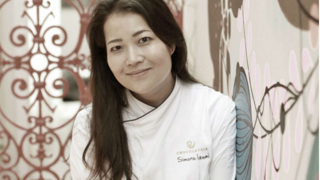 Na imagem vemos a Chef Simone Izumi ao centro com fundo de desenhos coloridos de flores. Simone tem traços orientais: cabelo liso preto e comprido e olhos castanhos puxados. Ela sorri de boca fechada. Simone veste seu uniforme de chef branco com o logo de sua confeitaria CHOCOLATRIA e seu nome estampados do lado esquerdo de seu peito em sua roupa. O logo é uma espiral da cor bege. Seu nome está escrito em marrom.