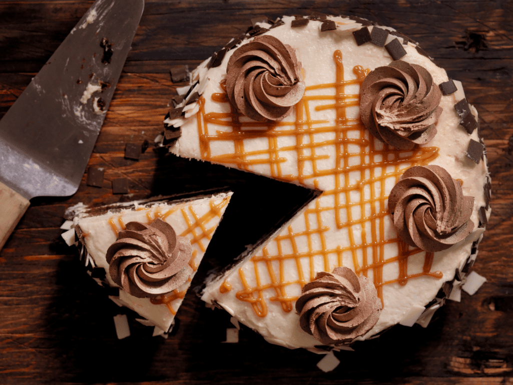 Bolo redondo visto de cima em uma mesa de madeira. O bolo tem uma cobertura branca e pequenas espirais de creme de chocolate em cima como decoração, além de calda de caramelo espalhada em traços verticais e horizontais.