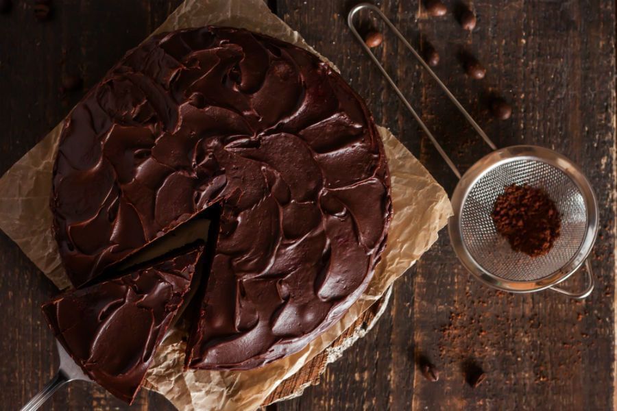 Bolo com cobertura de chocolate escura visto de cima. Ao lado vemos uma peneira de metal pequena com chocolate em dentro dela. O bolo está em cima de um papel da cor marrom em cima de uma mesa de madeira escura.