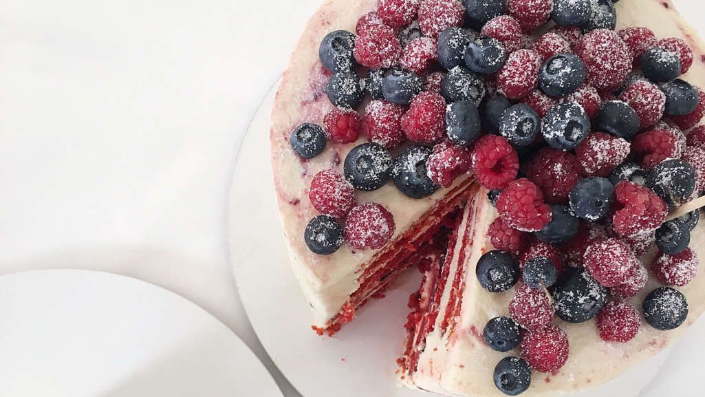Em uma mesa branca vemos um bolo com cobertura branca e com frutas vermelhas como amoras, framboesas e mirtilos em cima. O bolo falta uma fatia e sua massa é vermelha e recheio branco.