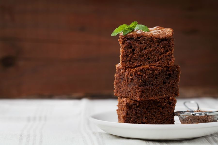 Em fundo de madeira, uma mesa coberta com um pano branco, há um prato branco e nele há três pedaços de bolo de chocolate empilhados, no pedaço de cima há folhas de hortelã decorando.