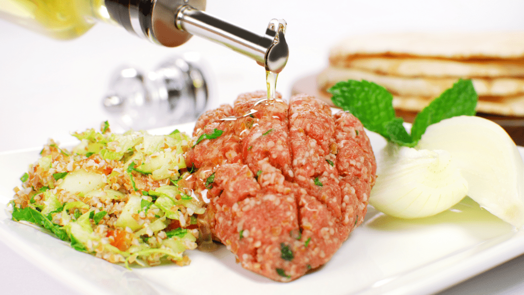 Em um prato branco há um kibe cru na cor vermelho vibrante com pequenas folhas verdes. Ao lado esquerdo temos tabule e do outro cebola fatiada e hortelã.