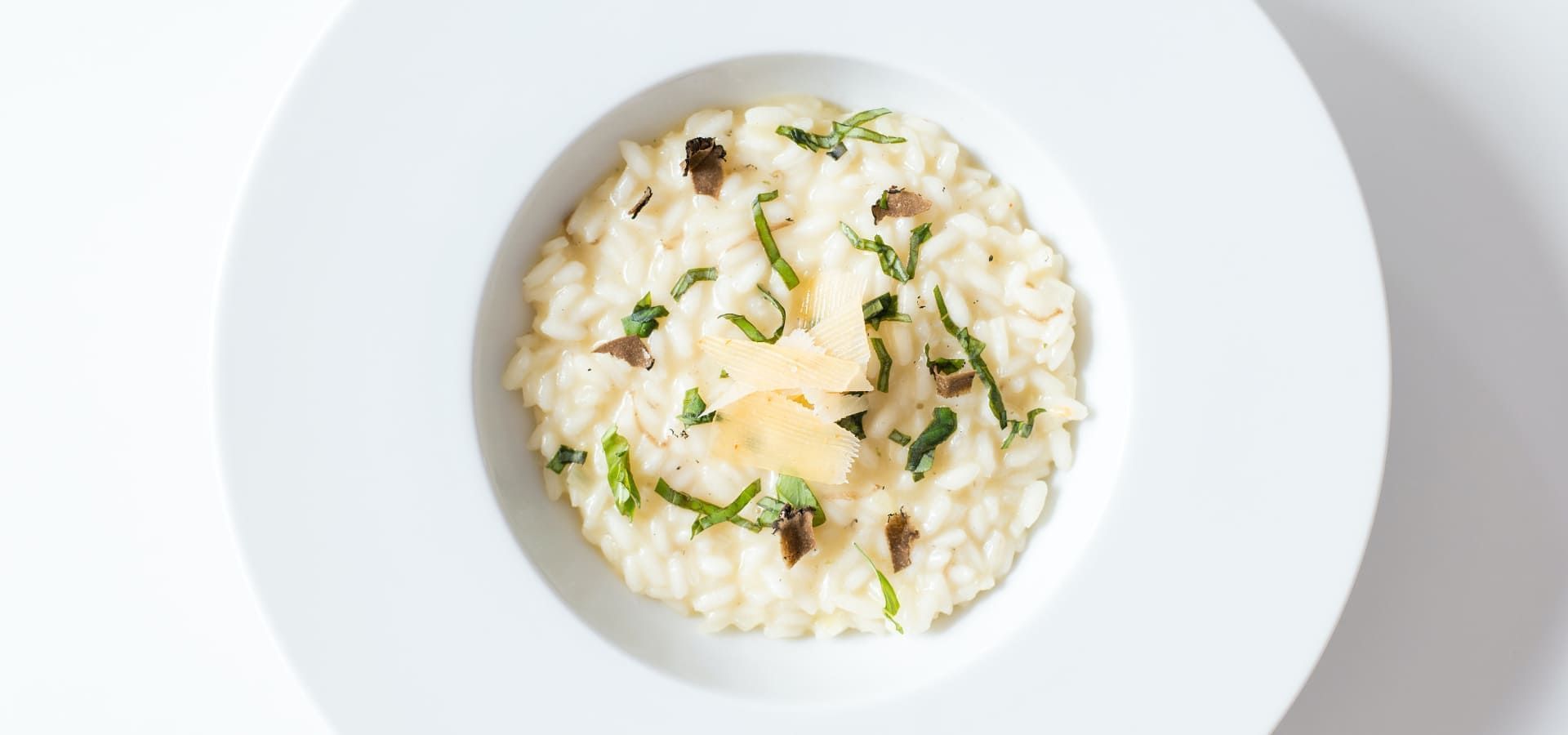 Vista de cima de prato branco em mesa branca com risoto de cor bege claro decorado com folhas verdes fatiadas e queijo.