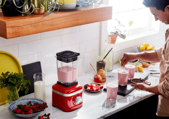 Black Friday 2021 - Liquidificador KitchenAid K150 batendo bebida de frutas vermelhas com leite em bancada branca com copos com bebida cor de rosa, leite e tigela de frutas vermelhas como morango.