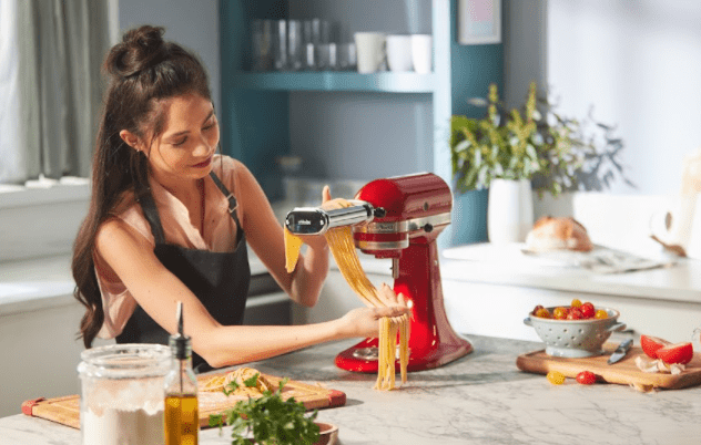 acessórios kitchenaid - Mulher com cabelos pretos manuazendo uma massa de macarrão sendo feita no pasta roller da KitchenAid em uma batedeira Artisan Vermelha.
